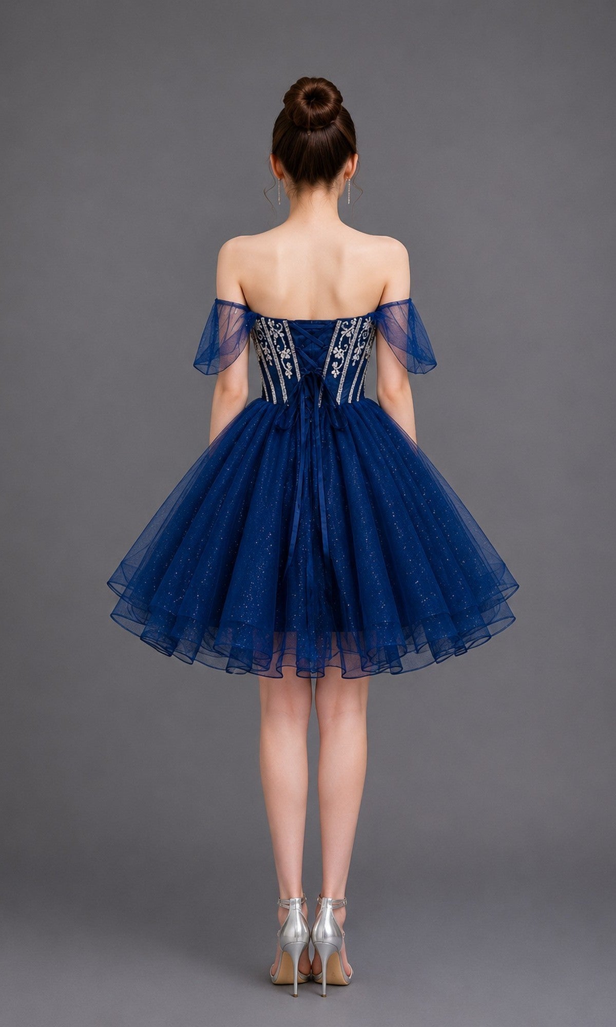 A woman stands with her back to the camera, wearing the Dancing Queen 3438 short homecoming dress—an off-shoulder ball-gown style that features a sparkly embroidered tulle skirt—paired with silver high heels against a plain gray background.